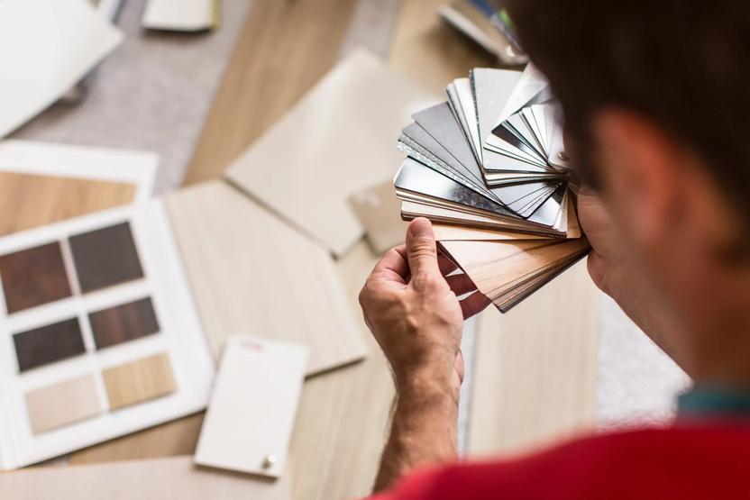 Nahaufnahme einer Person, die Farbmuster für Holzoberflächen in der Hand hält, umgeben von weiteren Materialien zur Auswahl.