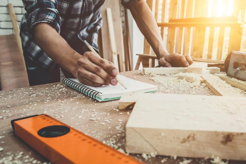 Handwerker notiert Ideen in einem Block auf einem Tisch, umgeben von Holzspänen und einem Winkelmaß.