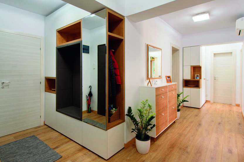 Heller Flur mit Holzregalen, Spiegel, Sideboard und Pflanzen in Töpfen. Eine Tür führt in einen weiteren Raum.