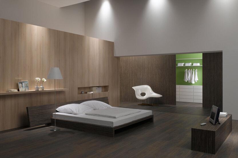 Schlafzimmerimpression mit Holzverkleidung an Wand Schlafzimmerimpression von Bett, Nachtisch und Sideboard mit Holzverkleidung an Wand und indirektert Beleuchtung
